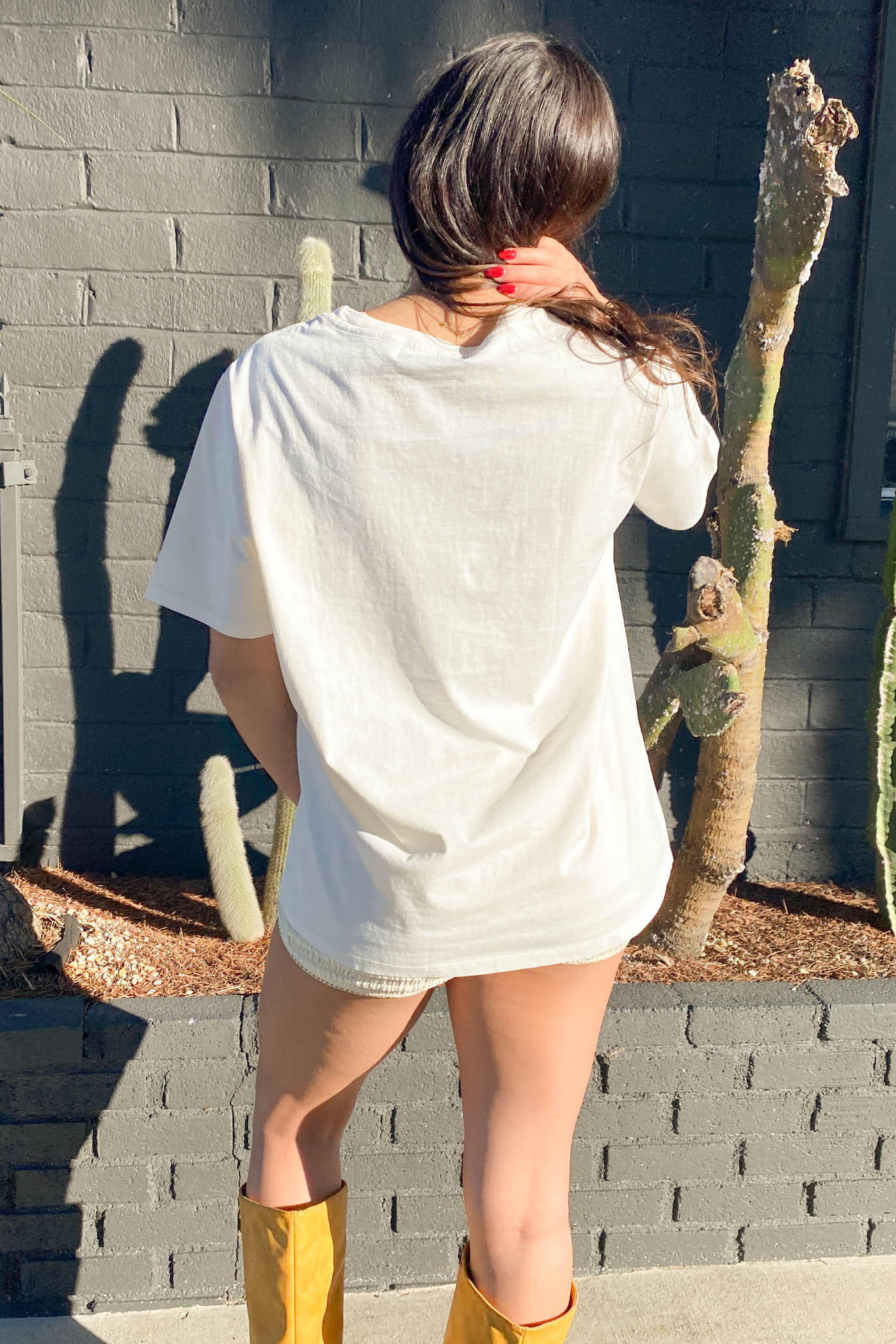 Person wearing a white oversized shirt and yellow boots standing against a gray brick wall with cacti.