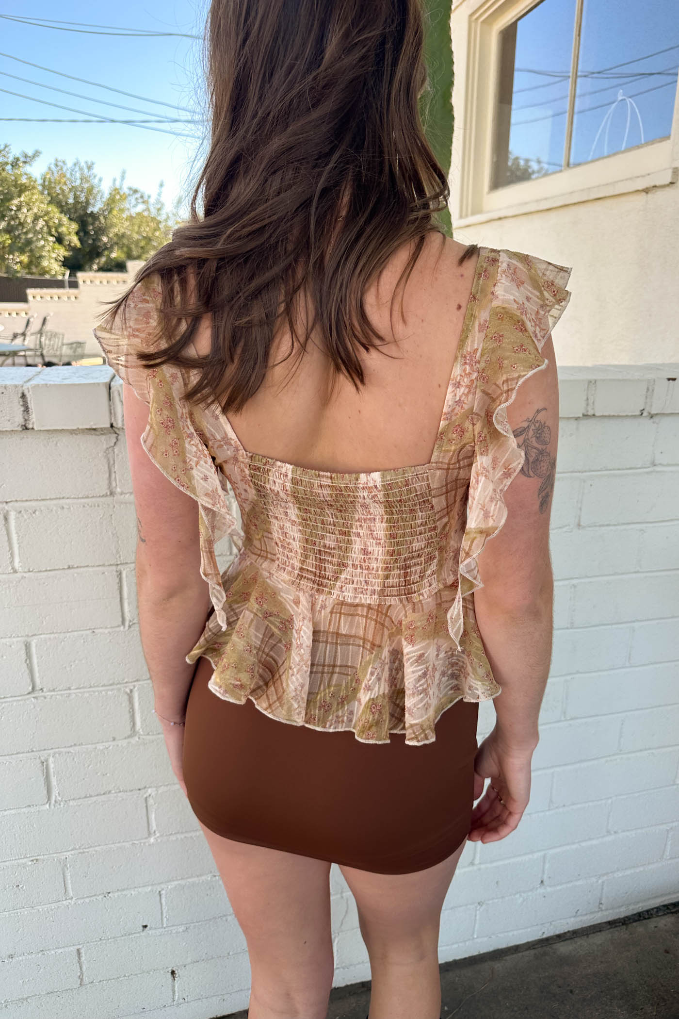 Woman wearing a patterned top and brown shorts standing against a white brick wall.