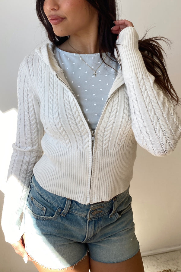 Woman wearing a white cable knit cardigan over a light blue polka dot shirt and denim shorts.