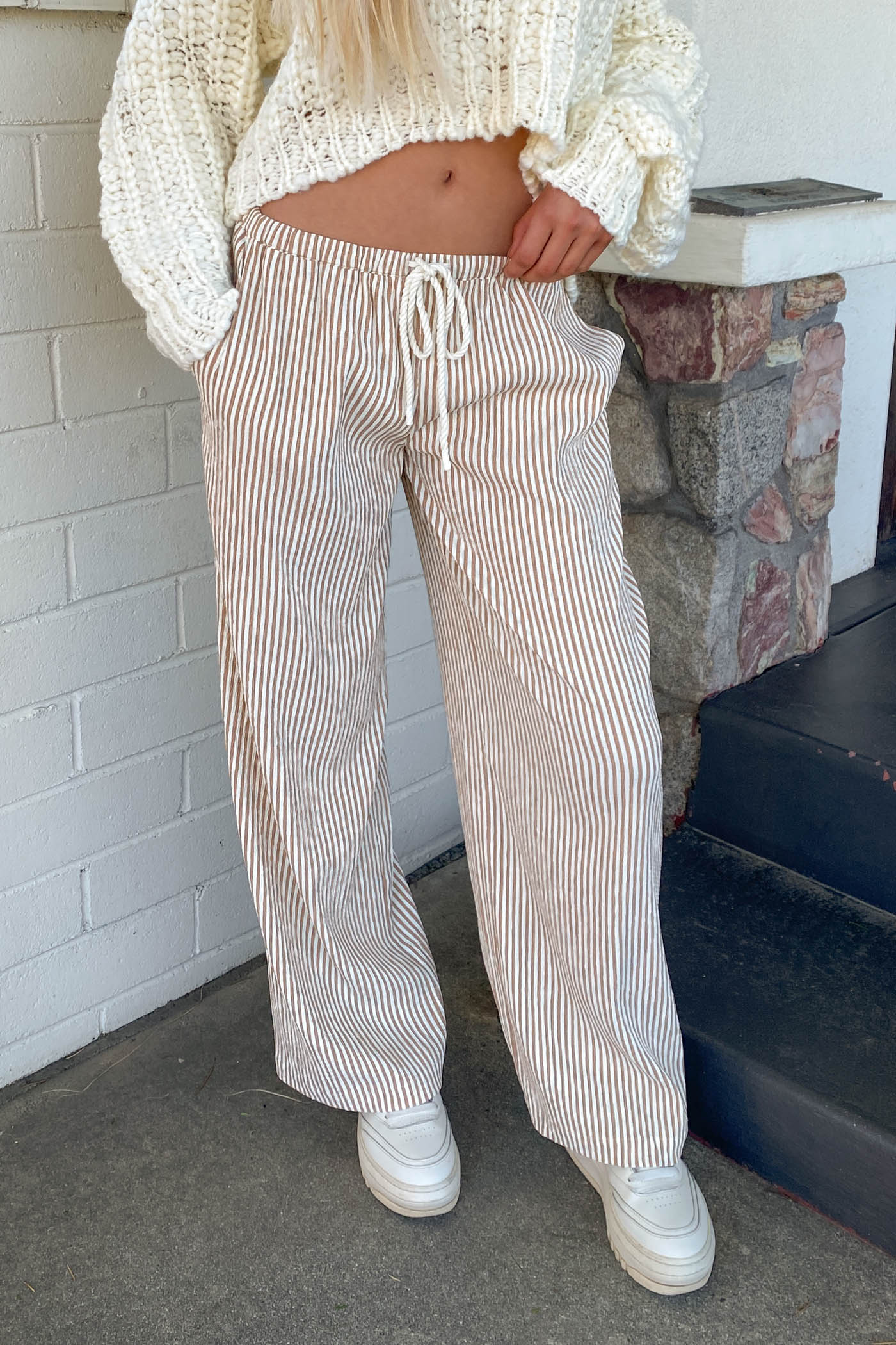 Person wearing a white knit sweater and striped pants standing against a brick wall.