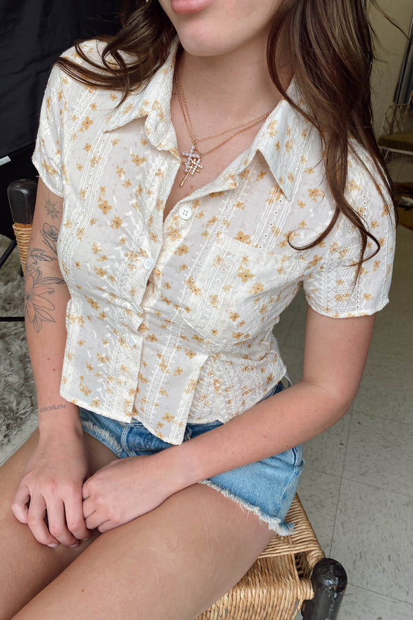 Person wearing a white floral blouse and denim shorts sitting on a chair.