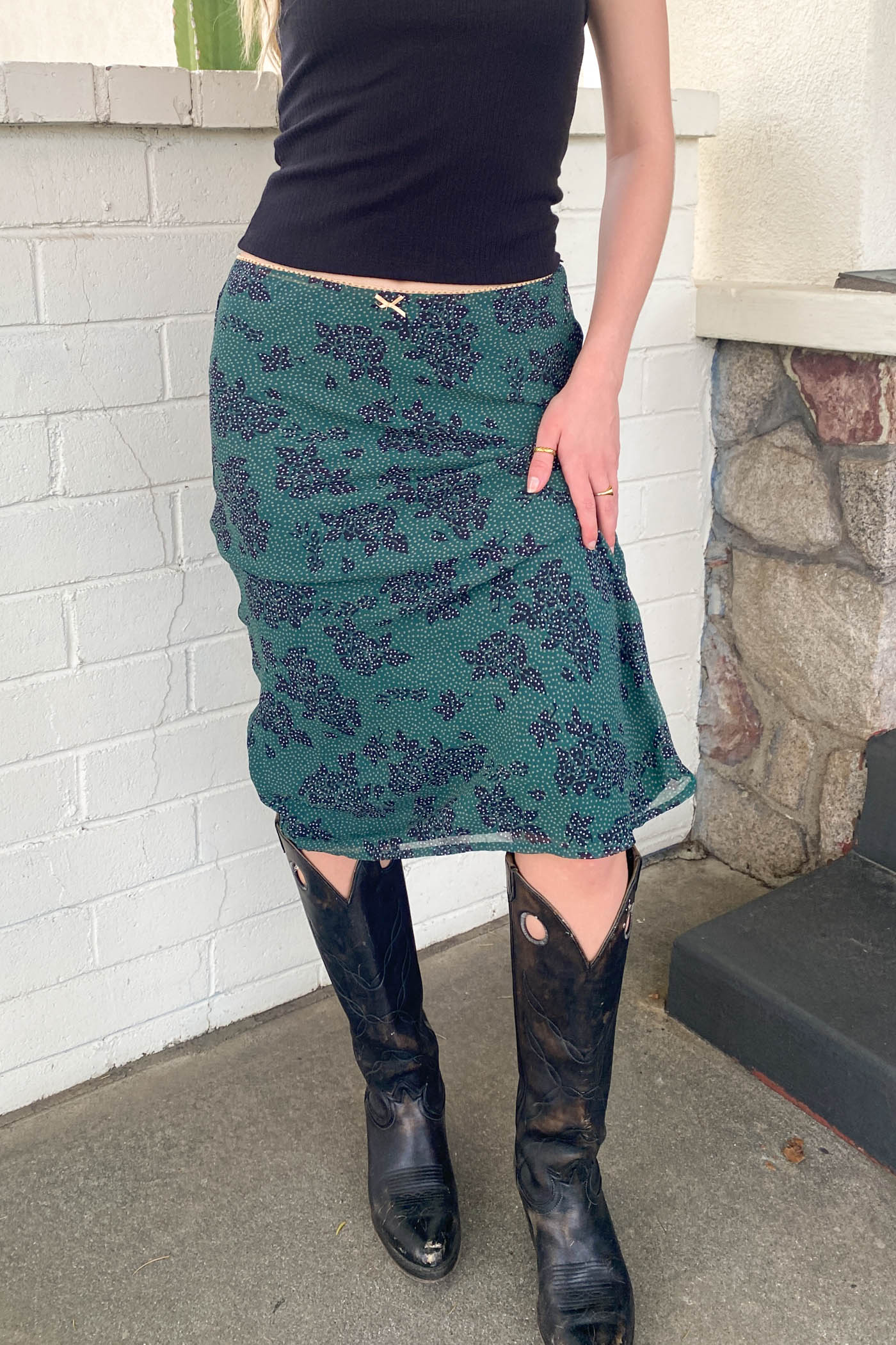Person wearing a green patterned skirt and black boots in front of a white brick wall.
