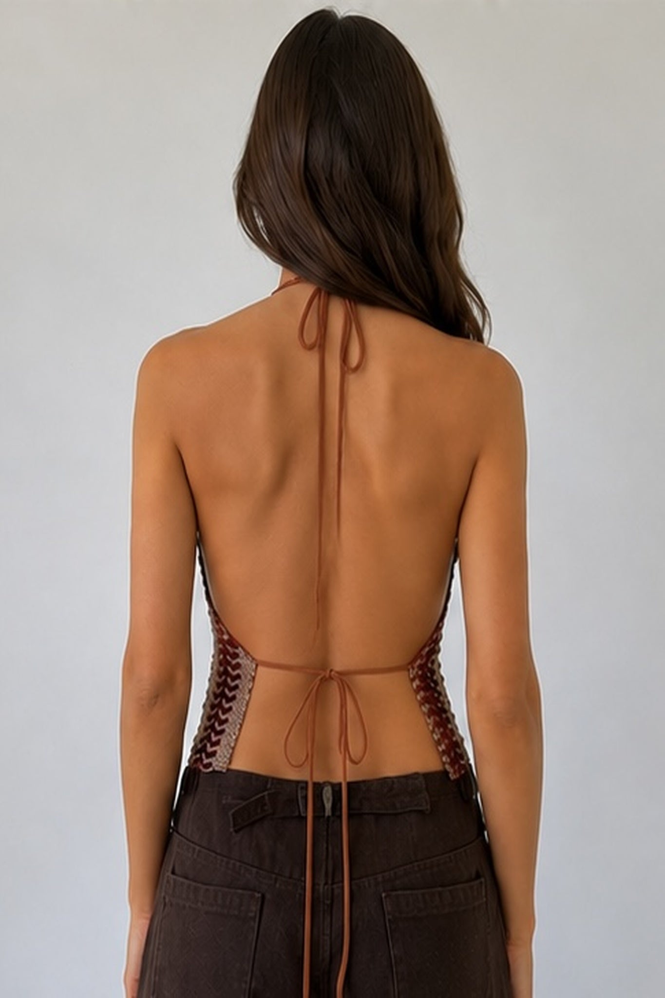 A woman viewed from the back wearing a brown lace-up halter top and dark brown pants against a plain light gray background.