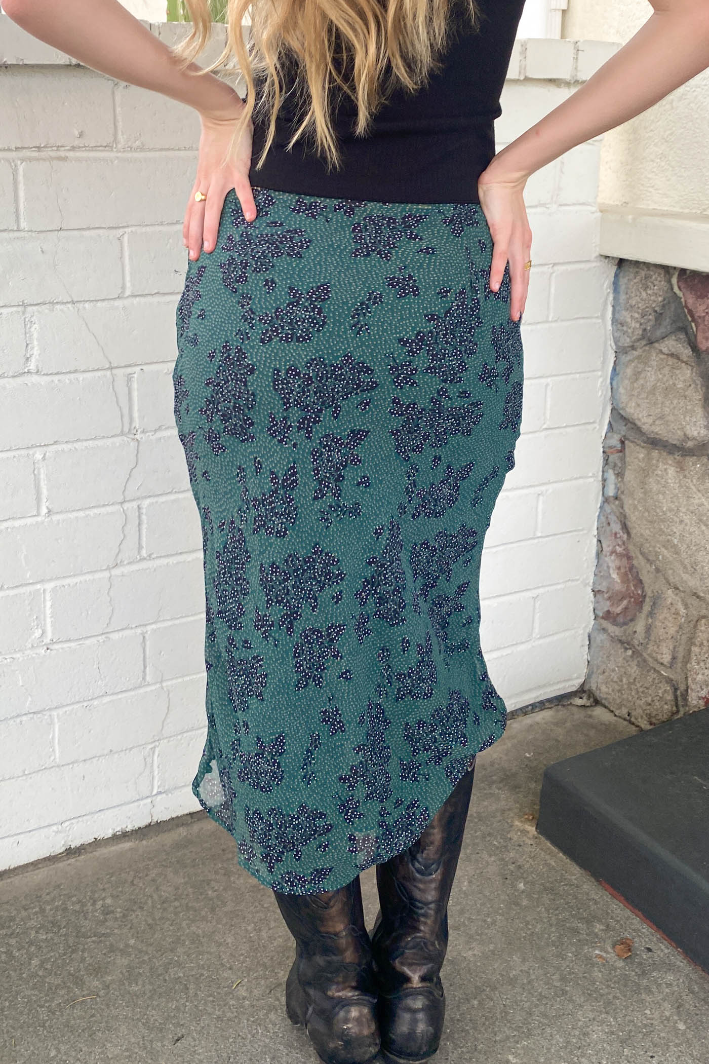 Person wearing a green patterned skirt with black boots against a white brick wall.