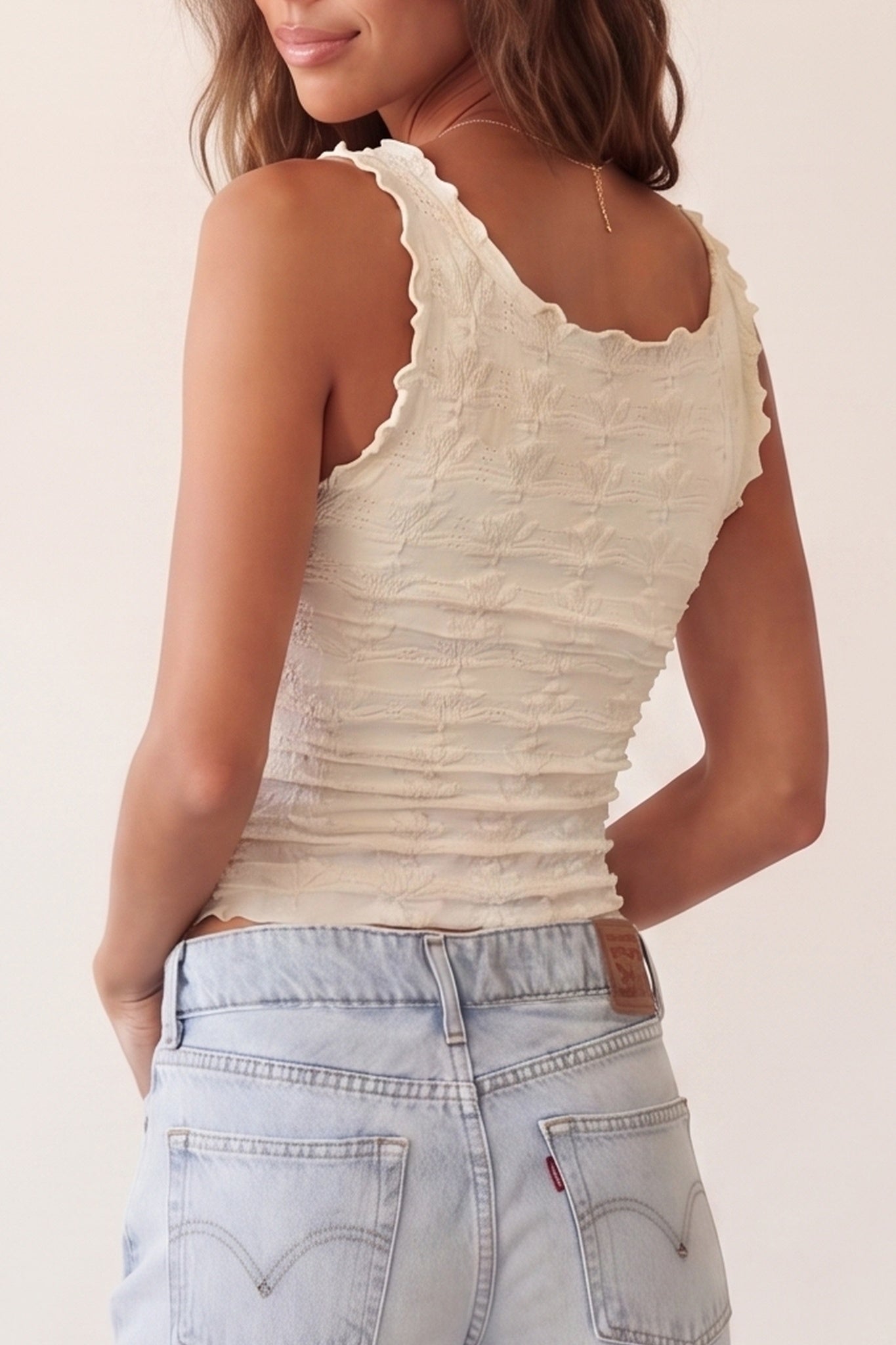 A woman wearing a cream-colored, textured sleeveless top with a scalloped neckline, paired with light-wash blue jeans, shown from a back-three-quarter view against a plain light beige background.