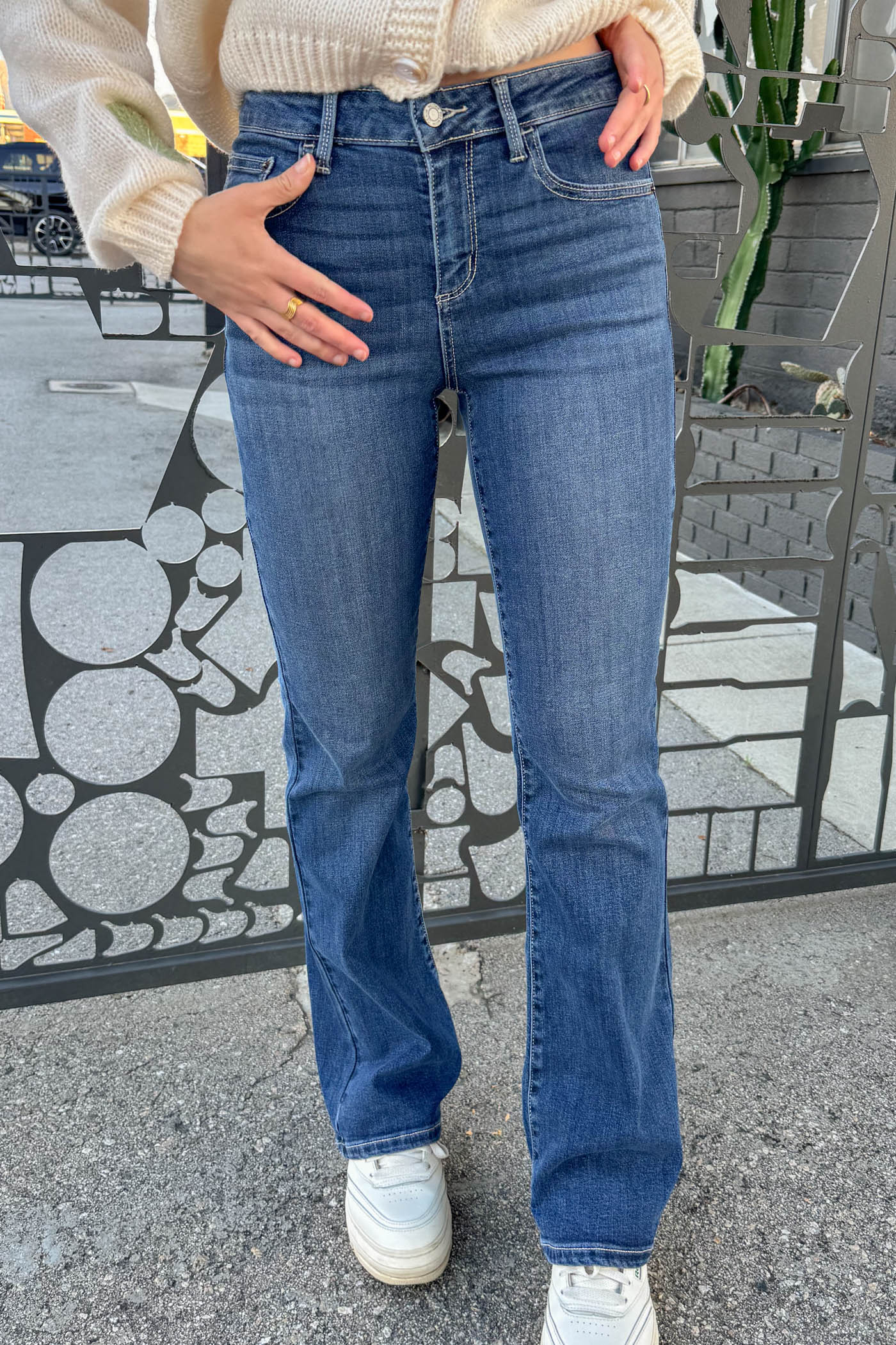 Person wearing blue jeans and a light-colored sweater standing on a street.