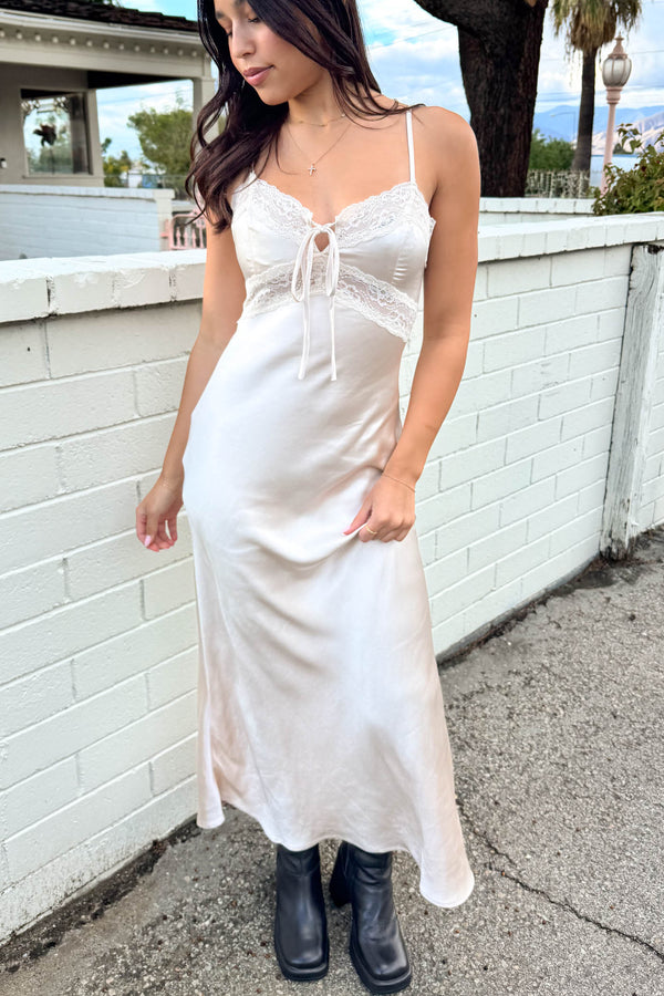 Woman wearing a white slip dress with lace details against a white wall.